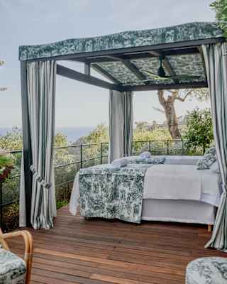 A gazebo shades two beds with grey Toile de Jouy fabric at Le Jardin des Rêves di Dior with a deck overlooking hills and sea.