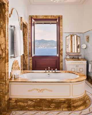 Looking through a white bathroom with ochre marble details, in door fames and a luxury bath, with an open, sea-view window.