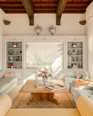 A bookcase, vintage sofa, two blue armchairs and yellow loveseat border a large coffee table beneath a beamed suite ceiling.
