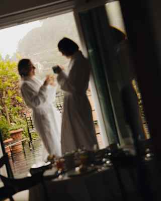 Angled image of two female guests in bath robes standing in a suite doorway, drinking tea, looking out onto misty gardens.