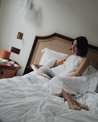 Angled image of a dark-haired woman in loose white clothing, reading as she reclines on a rumpled bed in her suite.