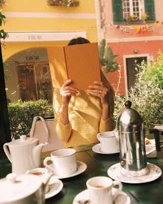 A woman in a yellow top holds an orange menu book in front of her face, seen over a table covered with coffee and teacups.