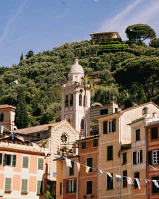 A verdant hill with a hilltop residence rises above the roofscape of Portofino, with gentle gables and pastel hues.