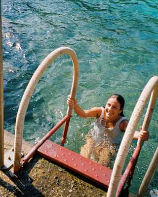 A smiling woman in a white swimming costume climbs metal steps from the clear sea up to a concrete platform, seen from above.