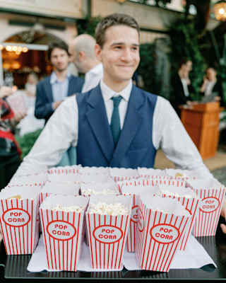 A staff member dressed as an usher with a navy waistcoat and teal tie holds a large tray of red-striped popcorn buckets.