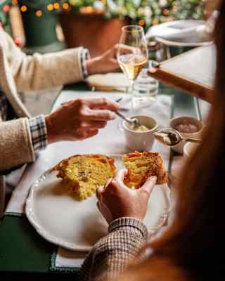 View over the shoulder of a diner who lifts a slice of panettone from a plate of two, as a companion stirs coffee.