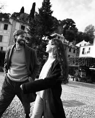 A man and woman walk together outdoors, smiling at each other. Trees and buildings are in the background. The photo is black and white.