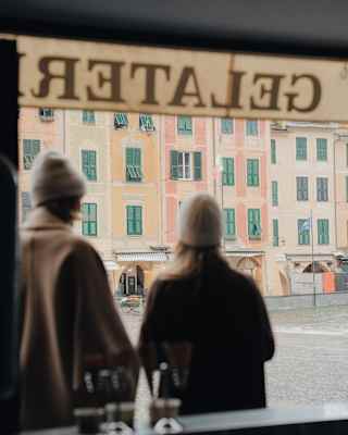 A couple in coats and hats gaze at Portofino's pastel edifices, seen from behind through the window of Gelateria San Giorgio.