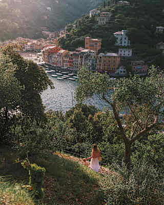 A woman in a white dress stands on a hillside path, overlooking a coastal village with colourful buildings and boats moored by the water, surrounded by lush green trees.