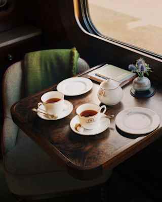 Two cups of tea sit on saucers atop a polished wooden train table with a teapot, china plates and a small vase of flowers.