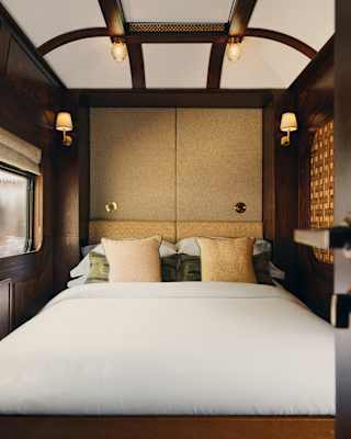 A double bed fills the polished wood cabin, with white linen, cream cushions and glowing wall lamps, seen through an open door.
