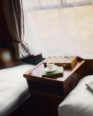 A cup of tea with a saucer rests on a green hard-backed book along with a folding chess board on a table between twin beds.
