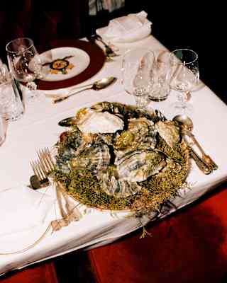 Looking down on a big plate of oysters in the shell, arranged dramatically on a moss bed, at a table with red velvet chairs.