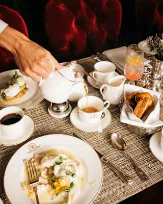 Close-up of the hand of a waiter as he pours tea from a pot into a cup at a breakfast table with poached eggs and pastries.
