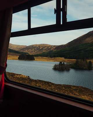 The train passes a loch with a forested island and mountainous backdrop in bronze and heather, seen through a window.