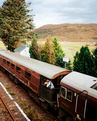 A man looks out from an observation deck as The Royal Scotsman waits at a rural station in the Highlands, seen from above.