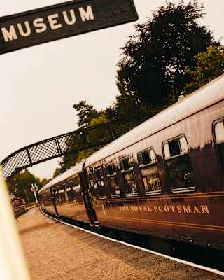 Angled image of The Royal Scotsman at a platform with a pedestrian bridge, seen from below a perpendicular sign for a museum.