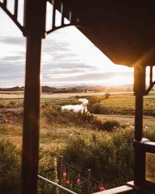A silver river snakes through fields as the sun glows on the horizon, seen from the Observation Car deck, framed by railings.