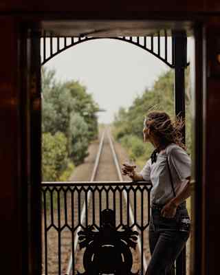 Wind flicks the long hair of a woman as she rests on the deck railings with a drink and watches the trailing landscape.