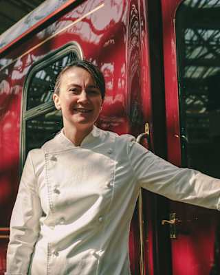 Chef Lorna McNee stands by the cherry exterior of The Royal Scotsman with one hand on a door handle, in a low-angle close-up.