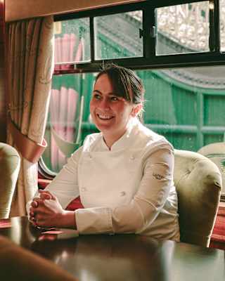 Chef Lorna McNee, wearing chef whites, sits behind a table inside a restaurant car with her hands clasped in front of her, smiling.