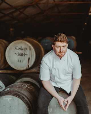 Chef Mark Donald, with red hair and beard, sits on a barrel in the Glenturret Distillery wearing chef whites and dark trousers.