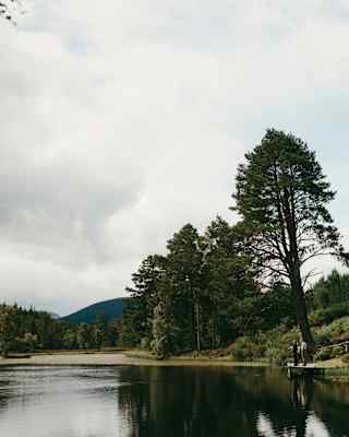 Seen across the water, two people stand on a small wooden deck between a grassy hill and a serene lake in the great outdoors.