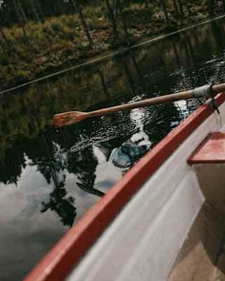 A paddle hovers above still lake waters in an angled image of a wooden oar resting on the side of a red-lipped rowing boat.
