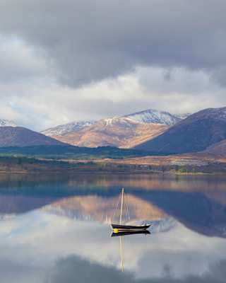 Two sailing boats sit on the glasslike waters of Loch Eil which mirror the surrounds of green fields and snow-capped mountains.
