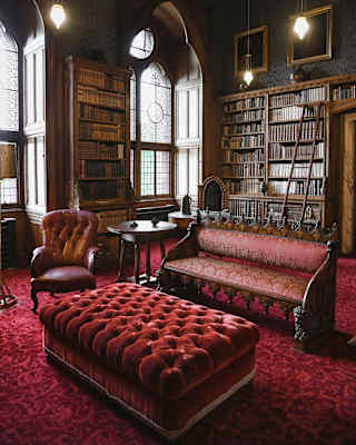 Two tall arched windows draw light into a library with wooden book shelves and a bench, armchair and pouffe in shades of red.