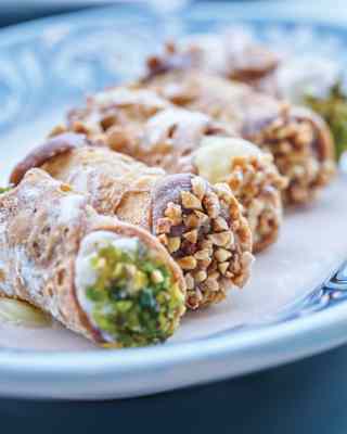 Close-up of five cannoli with pistachio, chocolate and vanilla cream, topped with nuts and served on a blue and white dish.