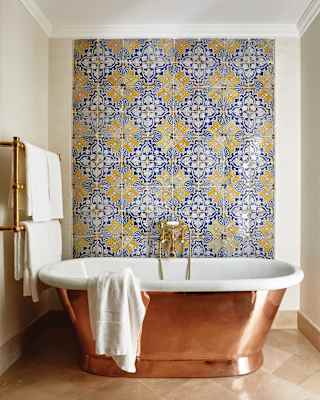 Blue and yellow geometric patterned tiles fill one wall. A free standing copper and ceramic bath reflects the marble floor