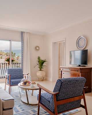 Two blue chairs face each other over a table of fruit in an room with balcony doors with views, and a bedroom to the right.