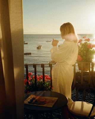 A woman in a white robe drinks coffee at her balcony rails gazing at boats on the sea, seen from behind in soft, honeyed light.
