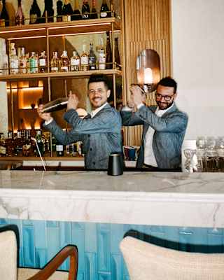 Two barmen, both with dark hair, beards and grey suits, one with glasses, smile as they shake cocktail shakers behind a bar.