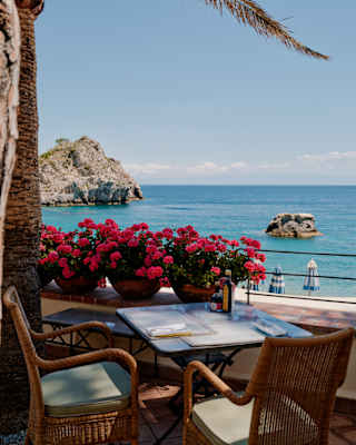A seaside terrace with two wicker chairs, a table set for two, vibrant pink flowers, and a scenic view of the sea, rocky islands, and clear blue sky.
