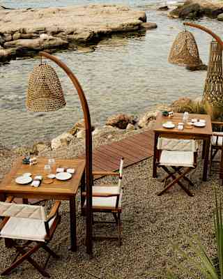 On a gravel bank by a coastal watery inlet, two Brizza dining tables with chairs and tall wicker standing lamps await guests.