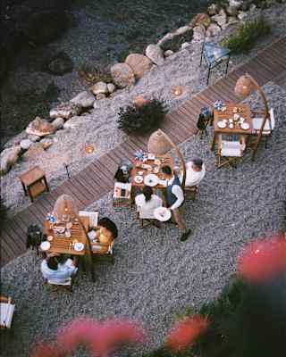 Aerial view of people dining at wooden tables on a gravel terrace near water, surrounded by rocks and plants, with warm ambient lighting.