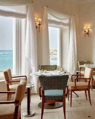 A long row of tables with chairs in pastel hues hug the wall of windows looking out on to Mazzarò Bay, in an airy dining room.