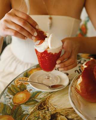 A blonde woman in a white dress, seen from the shoulders down, dips brioche into a strawberry granita at a café table.