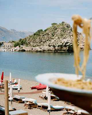 In soft-focus close-up, a diner raises spaghetti from a plate against a crisp backdrop of Mazzarò beach and its headland.