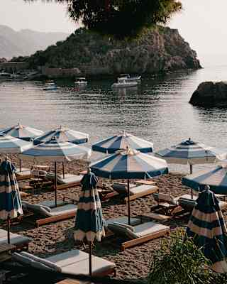 A shot of blue and white parasols on Mazzarò Bay beach at sundown, with views across still waters and boats to the headland.