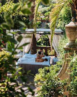 Two guests, face down on treatment beds and covered with towels, receive massages in the shade of the plant-filled terrace.