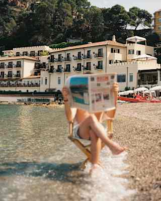 A man sits on a chair in the shallows, holding a newspaper in front of his face, seen in soft-focus on the hotel's beach.