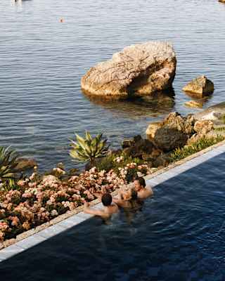 Three swimmers rest at the edge of the pool, gazing out past a small rock garden beach and nearby sea stacks to the ocean.