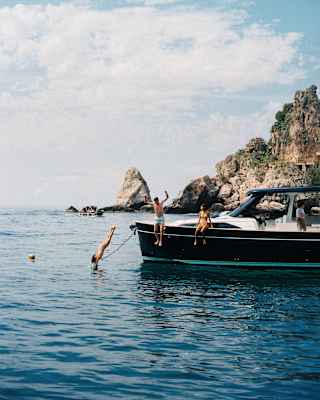 Action shot as a woman dives and a man leaps from a pleasure boat into the sea, watched by a woman who sits on the edge.