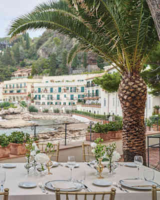 A long table is exquisitely set for lunch on the dining terrace amid palm trees and looking across the Bay at the sweep of hotel