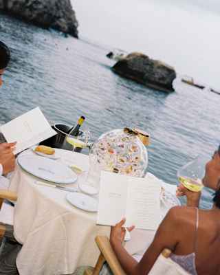 Angled shot of a couple, mostly off-camera, as they peruse menus at a waterfront table with wine and a glass globe ornament.