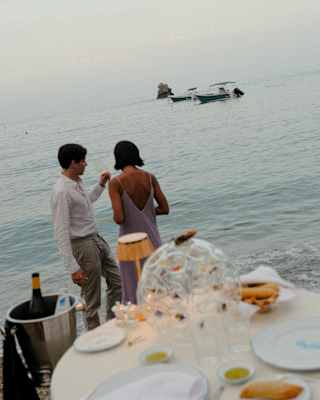 A couple stand on the shoreline facing the sea and boats, sharing an evening drink, seen at an angle over a candle-lit table.