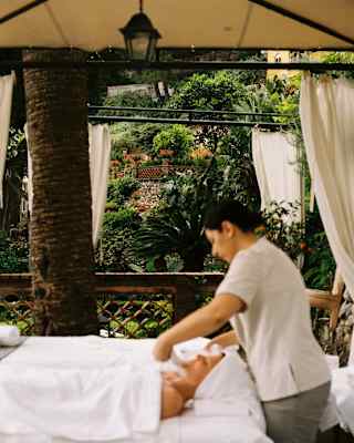 A therapist wraps a towel around the face of a woman who lies on an outdoor spa table beneath a white sheet, in soft-focus.
